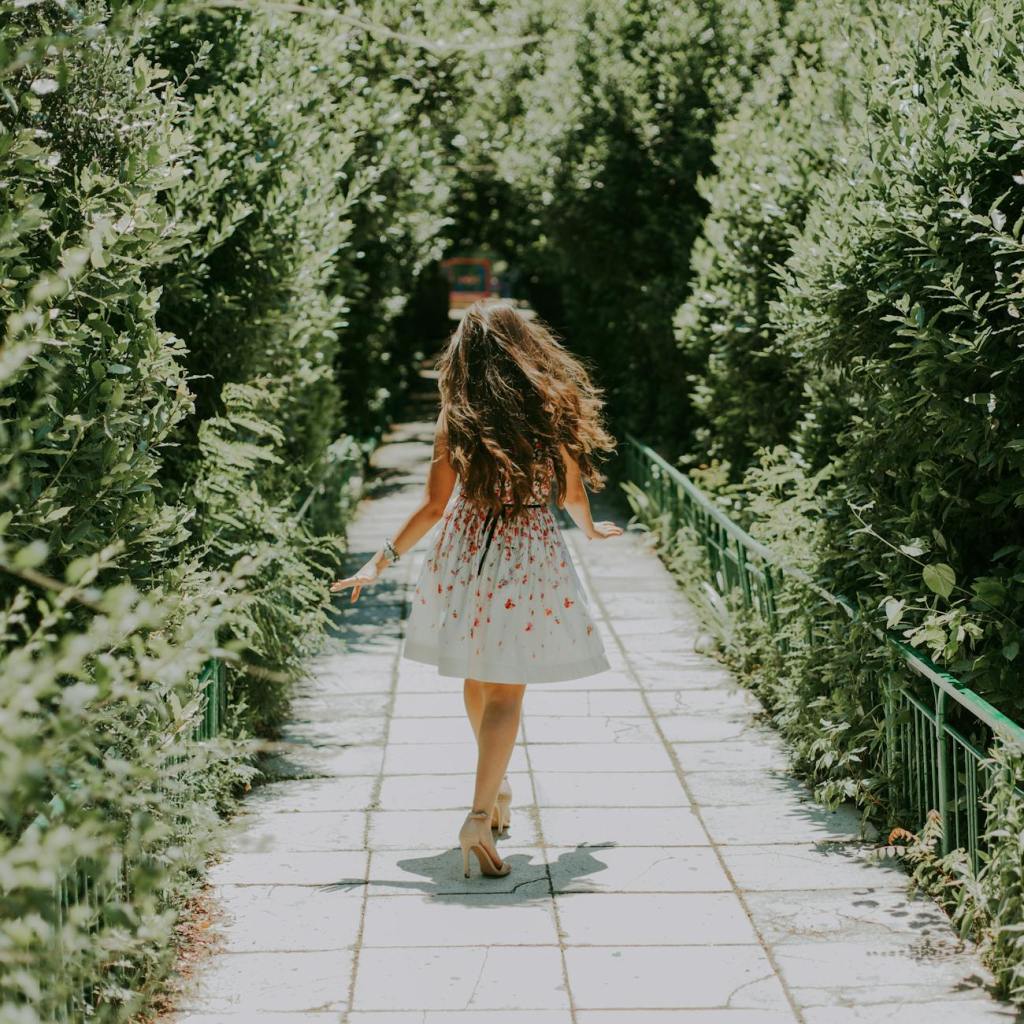 photography of woman in white and red floral midi dress walking on pathway